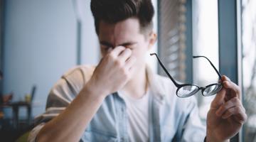 A young man holding glasses in one hand and touching his closed eyes with another hand