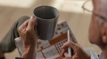 A senior man holding a mug, newspaper and a pen on his lap